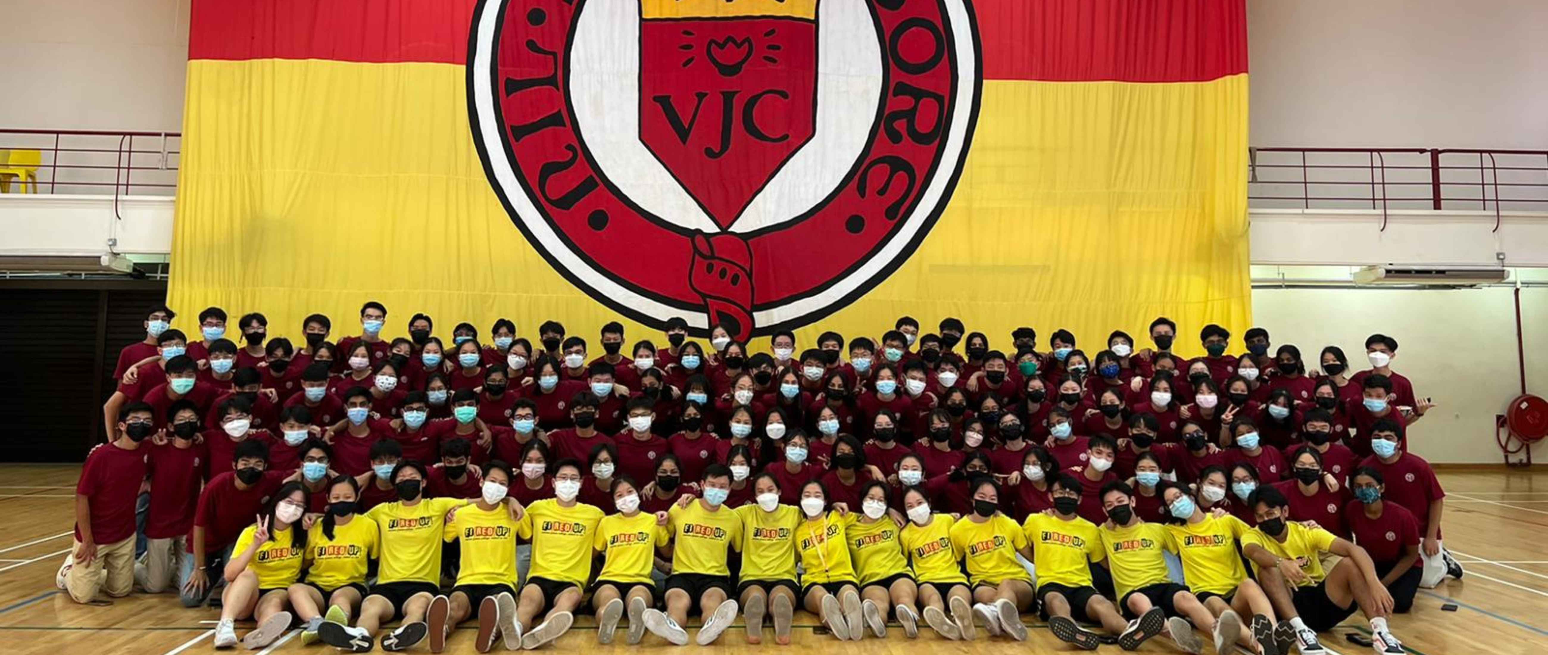 Large group of masked students in red and yellow shirts posing in a gym before a large VJC school flag.